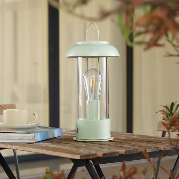 Tragbare LED-Laterne mit mintgrünem Gehäuse und Glaszylinder auf einem hölzernen Gartentisch neben einer Kaffeetasse und einem Buch.