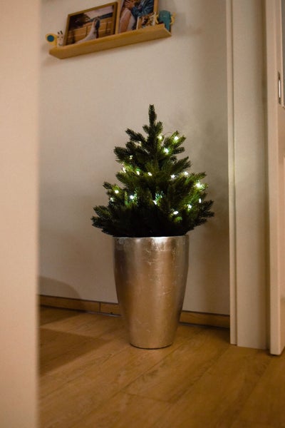 Künstlicher Weihnachtsbaum mit LED Lichterkette in einem silbernen Übertopf auf einem Boden in Holzoptik.