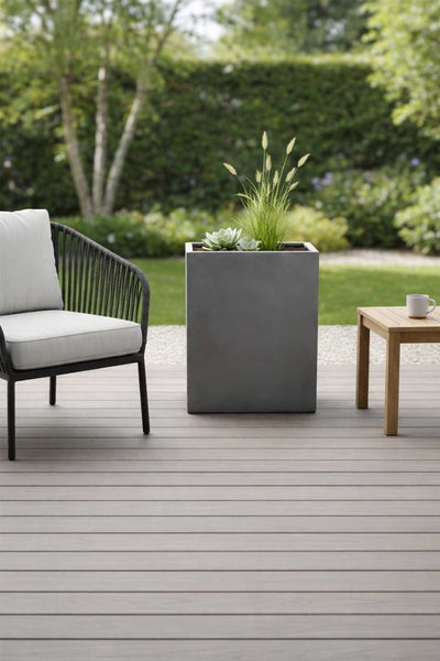 Moderner grauer Pflanzkübel auf einer Holzterrasse neben einem Gartensessel und einem Beistelltisch in einem grünen Garten.
