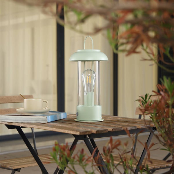 Moderne hellgrüne LED-Tischleuchte in Laternenform auf einem Holztisch im Freien neben einer Tasse und einem Buch.
