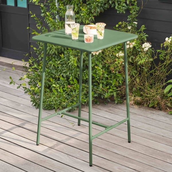 Quadratischer Bartisch aus Metall in Grün auf einer Holzterrasse mit Getränken und Pflanzen im Hintergrund.