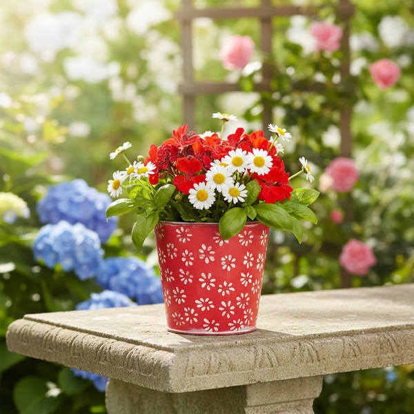 Roter Blumentopf mit weißen Blumenmustern, bepflanzt mit roten Geranien und weißen Gänseblümchen, auf einer Steinbank in einem hellen Garten.