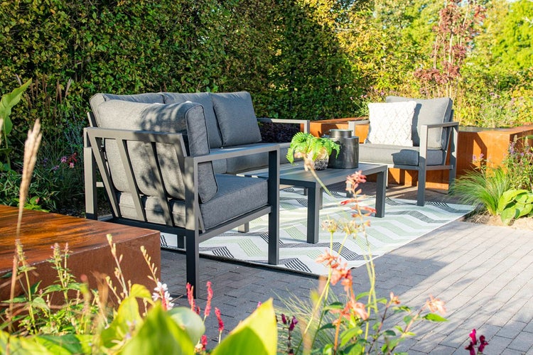 Modernes Garten-Lounge-Set aus grauem Metall mit Sofa, Sessel und Couchtisch auf einem Außenteppich in einem grünen Garten.