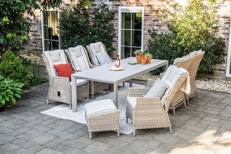 Gartenmöbel-Set mit großem Esstisch, Polyrattan-Sesseln inklusive Sitzkissen und einem Hocker auf einer gepflasterten Terrasse vor einer Ziegelwand.
