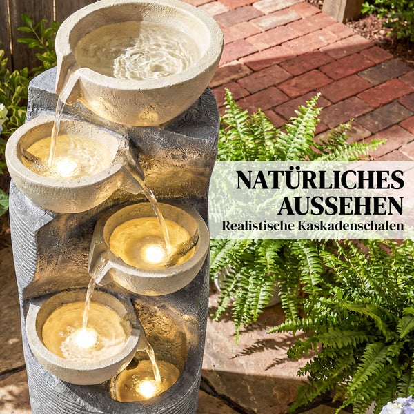 Gartenbrunnen in natürlicher Steinoptik mit Kaskadenschalen und integrierter Beleuchtung in einem hellen Garten mit Pflanzen und Ziegelweg.
