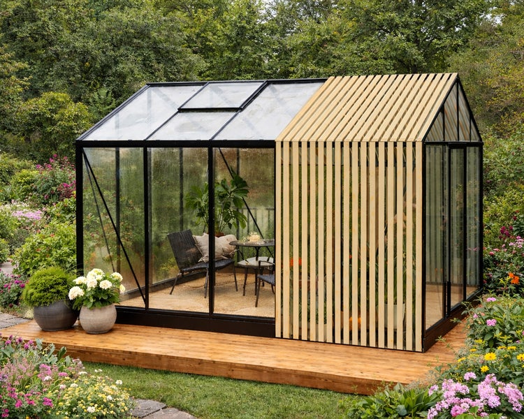 Modernes Gewächshaus mit schwarzem Rahmen, Glaswänden und Holzlamellen auf einer Holzterrasse im Garten, mit Sitzgruppe im Inneren.