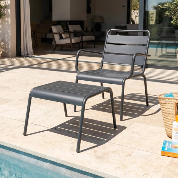Anthrazitfarbener Loungesessel und passender Hocker aus Metall auf einer hellen Steinterrasse am Pool.