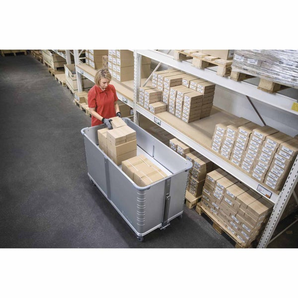 Eine Frau in einem organisierten Lagerhaus stapelt Pappkartons in einen großen fahrbaren Transportbehälter aus Metall vor hohen Metallregalen.