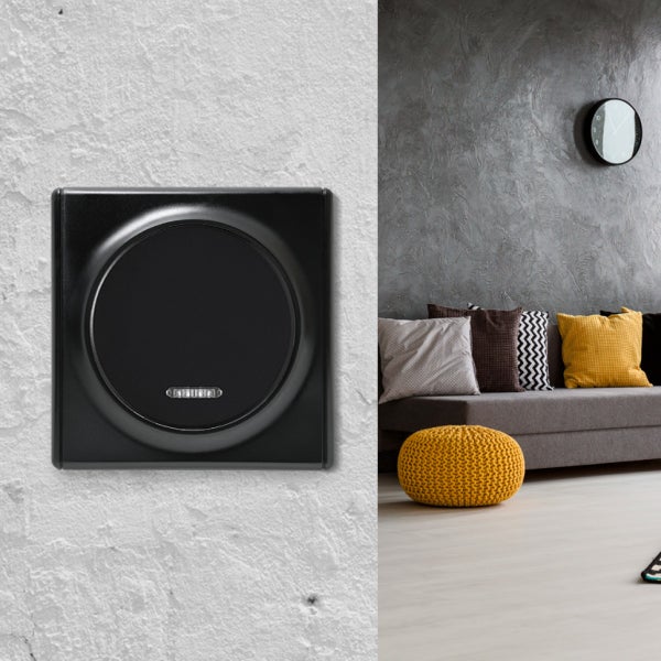 Ein moderner Lichtschalter mit rundem Bedienelement an einer hellen Wand, daneben ein Ausschnitt eines Wohnzimmers mit Sofa und Dekokissen.