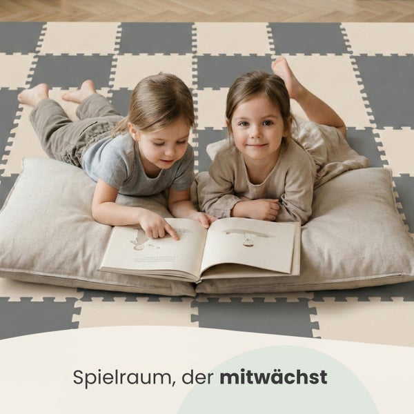 Zwei Kinder liegen auf einer grau-beigen Puzzle-Spielmatte aus Schaumstoff und lesen gemeinsam ein Buch auf Kissen.