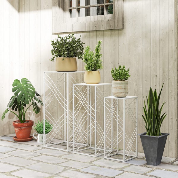 Drei weiße Blumenständer aus Metall in verschiedenen Höhen mit Gittermuster auf einer Terrasse mit Holzwand, Steinboden und diversen Pflanzen.