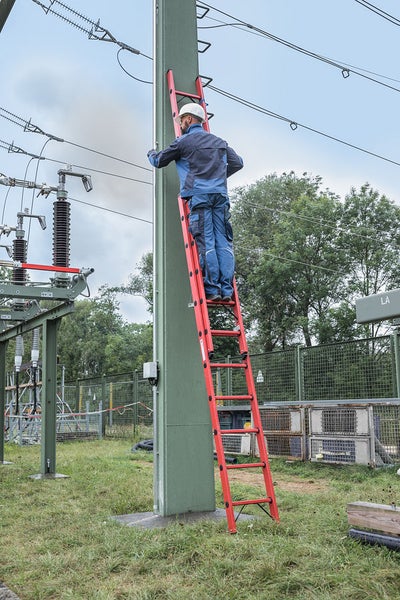 Ein Mann in Arbeitskleidung und Schutzhelm steht auf einer Anlegeleiter an einem Strommast.