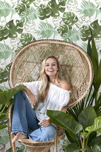 Eine Frau sitzt in einem Rattansessel vor einer Tapete mit Pflanzenmotiven.