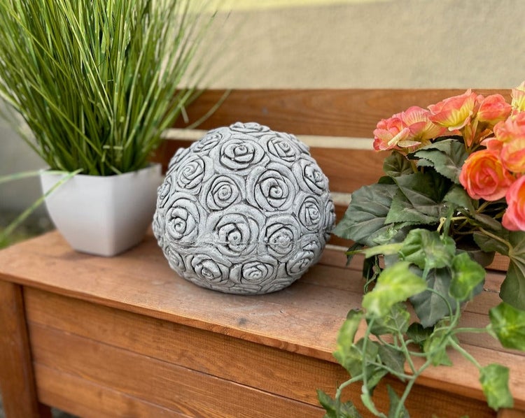 Graue Deko-Gartenkugel aus Stein mit Rosen-Relief auf einer Holzbank neben Topfpflanzen.