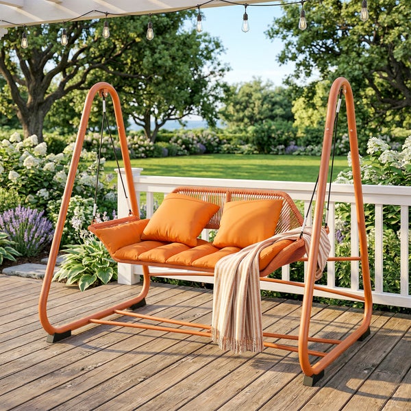 Moderne orangefarbene Gartenschaukel auf einer Holzterrasse mit Kissen und Decke vor einem weißen Zaun und Gartenhintergrund.