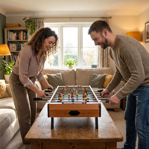 Ein Mann und eine Frau spielen mit einem Tischkicker aus Holz auf einem Couchtisch in einem hellen Wohnzimmer.