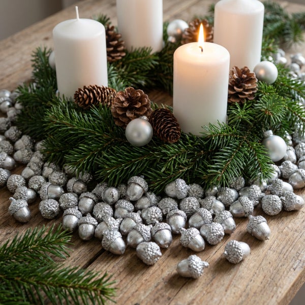 Adventskranz aus Tannenzweigen mit vier weißen Stumpenkerzen, Zapfen und silbernen Kugeln auf einem rustikalen Holztisch mit Dekoeicheln.