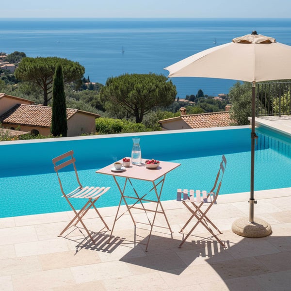 Ein modernes Bistroset aus Metall mit einem klappbaren Tisch und zwei Stühlen auf einer hellen Terrasse neben einem Swimmingpool mit Meerblick.
