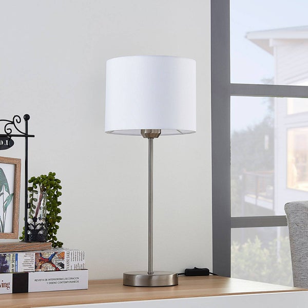 Moderne Tischleuchte mit weißem Stoffschirm und schlankem Metallfuß auf einem hellen Holztisch in einem hellen, organisierten Zimmer.