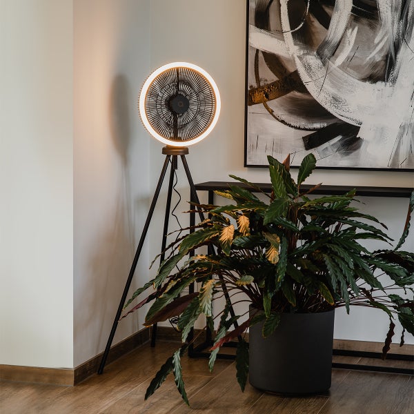 Moderner Stativ-Ventilator mit Lichtring in einem hellen Wohnraum neben einer großen Zimmerpflanze und einem Konsolentisch.