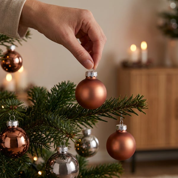 Hand platziert eine matte Weihnachtskugel am Weihnachtsbaum. Im Hintergrund ist eine wohnliche, weihnachtliche Dekoration zu sehen.