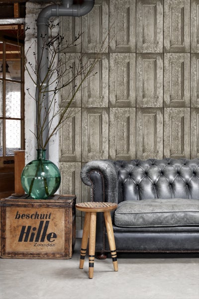 Wohnraum mit grauer Couch, Holzkiste, Holzhocker, grüner Glasvase und Tapete im Türenmuster