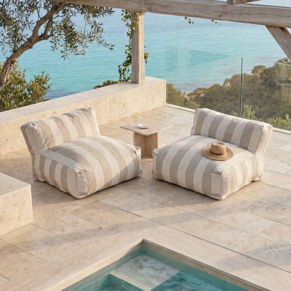 Zwei gestreifte Outdoor-Sitzsäcke auf einer hellen Steinterrasse mit Meerblick, Steintisch und Glasgeländer unter einer Holzpergola.