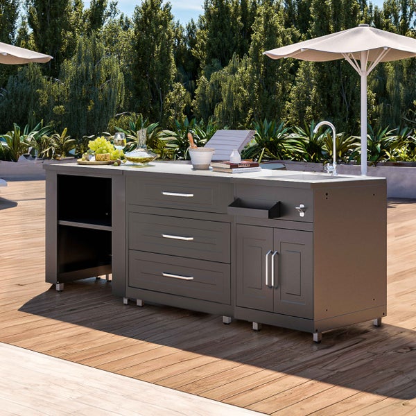 Graue Außenküche auf einer Holzterrasse mit Spülbecken, Armatur und drei Schubladen vor grünen Bäumen.