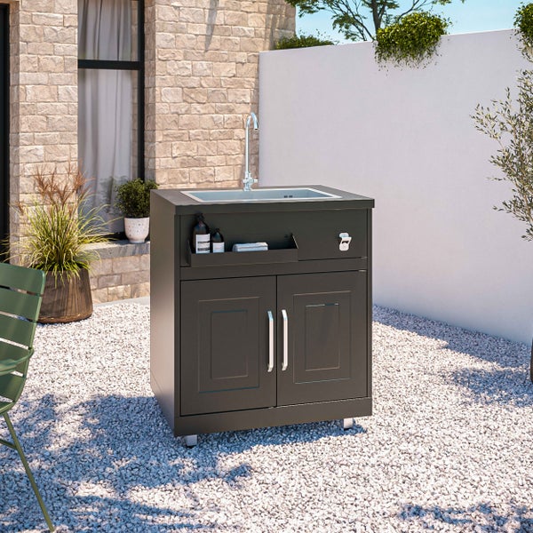 Moderne Außenküche in Anthrazit mit Edelstahlspüle und Unterschrank auf einer Kiesfläche im Garten neben einer Steinmauer.