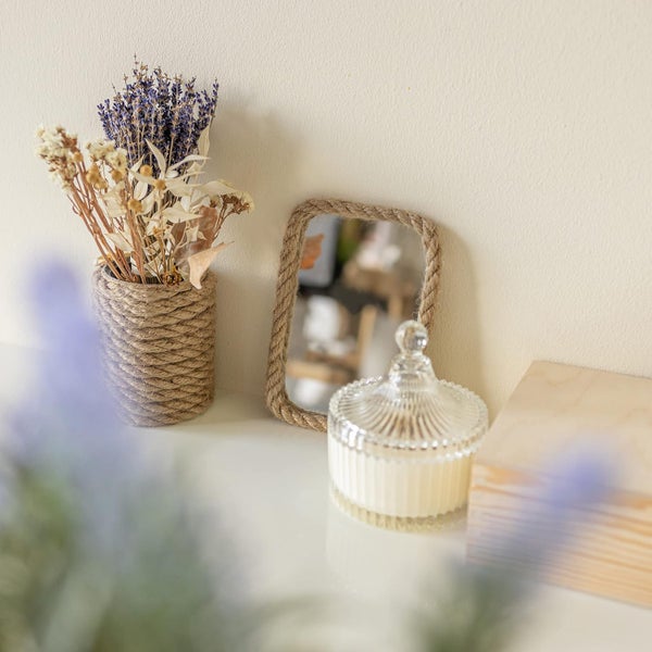 Kleiner rechteckiger Spiegel mit Seilrahmen, mit Seil umwickelte Vase mit Trockenblumen, geriffeltes Glasgefäß und Holzbox auf weißer Fläche.