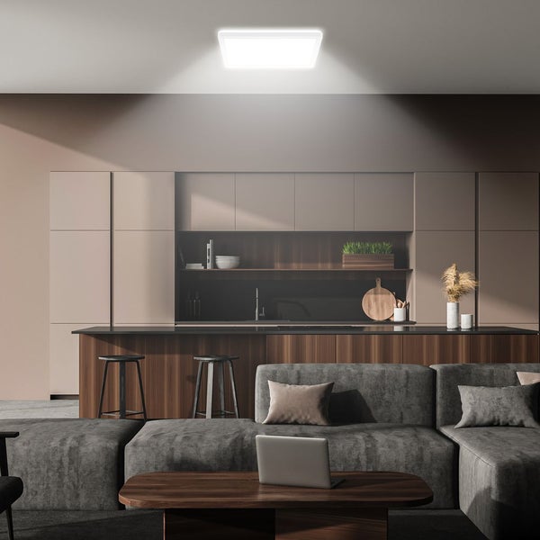 Quadratisches LED-Deckenpanel beleuchtet einen modernen, offenen Wohn- und Küchenbereich mit grauem Ecksofa und Kochinsel aus dunklem Holz.
