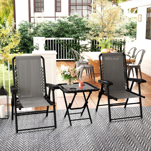 Zwei graue Gartenklappstühle und ein schwarzer Beistelltisch auf einer hellen Holzterrasse mit Pflanzen im Hintergrund.