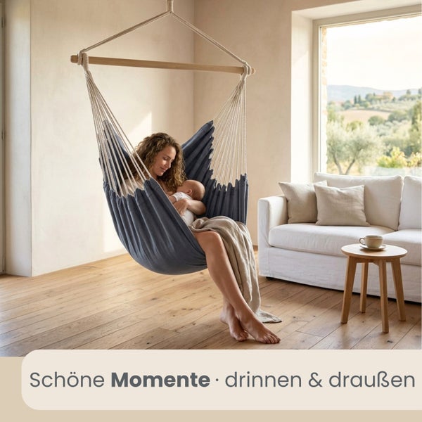 Frau mit Baby in einem blauen Hängesessel in einem hellen Wohnzimmer mit Holzboden, weißem Sofa und großem Fenster.