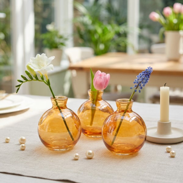 Drei kleine, runde Glasvasen in Bernsteinoptik auf einem Tisch mit Blumen und einer Kerze als dekoratives Arrangement.