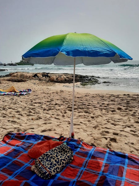 Sonnenschirm mit Farbverlauf von Grün zu Blau auf einem Sandstrand, im Vordergrund eine karierte Picknickdecke.