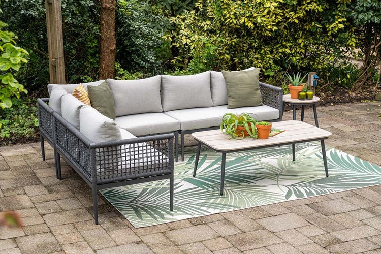 Garten-Lounge-Set mit Ecksofa, grauen Polstern, Seilgeflecht-Rahmen, Couchtisch und Teppich mit Palmenmuster auf einer Terrasse.