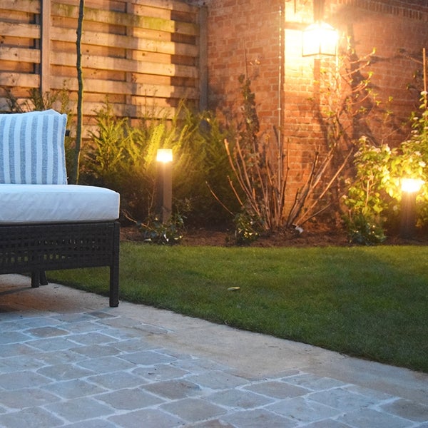 Beleuchteter Garten bei Nacht mit Pollerleuchten im Beet, Wandleuchte an der Ziegelwand und Gartenmöbeln auf einer Steinterrasse.