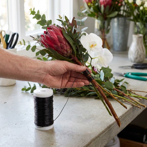 Eine Hand bindet einen Blumenstrauß aus Protea, Orchideen und Eukalyptuszweigen mit einem Wickeldraht auf einer Werkbank.