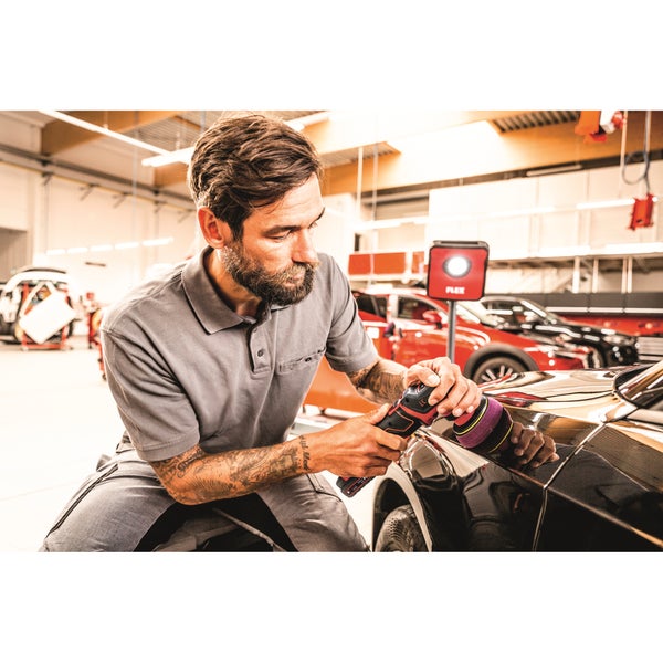 Ein Handwerker poliert mit einer akkubetriebenen Poliermaschine die Karosserie eines schwarzen Autos in einer hellen Werkstatt.