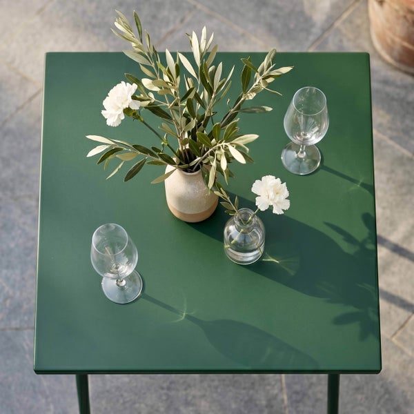 Quadratischer Gartentisch aus Metall auf einer Terrasse, dekoriert mit Vasen, Blumen und Weingläsern.