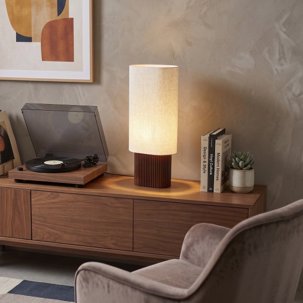 Moderne Tischleuchte mit zylindrischem Textilschirm auf geriffeltem Holzfuß, platziert auf einem Sideboard neben einem Plattenspieler.