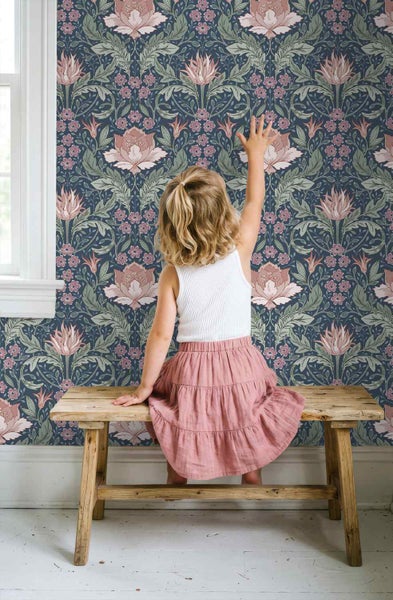 Ein Kind sitzt auf einer rustikalen Holzbank vor einer Wand mit floral gemusterter Tapete mit großen Blüten und Blättern.