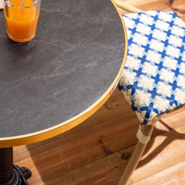 Runder Bistrotisch mit dunkler Steinoptik-Platte und Goldrand neben einem geflochtenen Bistrostuhl auf einer Holzterrasse.