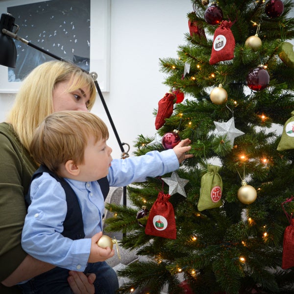 Eine Frau hält ein kleines Kind auf dem Schoß, das einen Weihnachtsbaum mit Kugeln, Sternen und Adventskalender Säckchen schmückt.
