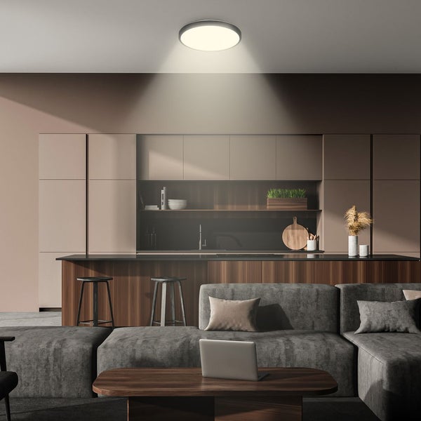 Runde LED-Deckenleuchte beleuchtet eine moderne, offene Küche und ein Wohnzimmer mit grauem Sofa und Holzmöbeln.