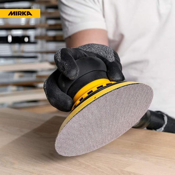 Eine Hand im Handschuh hält einen Mirka Exzenterschleifer beim Schleifen einer Holzoberfläche.