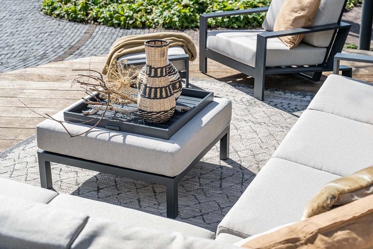 Moderne Gartenlounge mit Metallgestell, Polsterauflagen, Hocker mit Tablett und Außenteppich auf einer Terrasse.