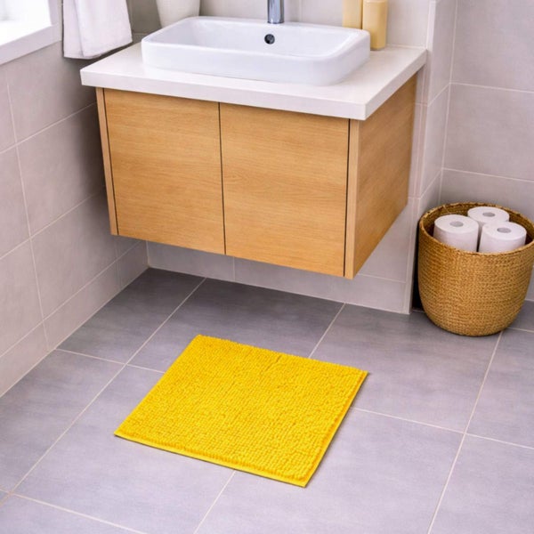 Gelbe Badematte auf Fliesenboden vor einem hölzernen Waschtisch mit weißem Waschbecken und einem Korb mit Toilettenpapier.