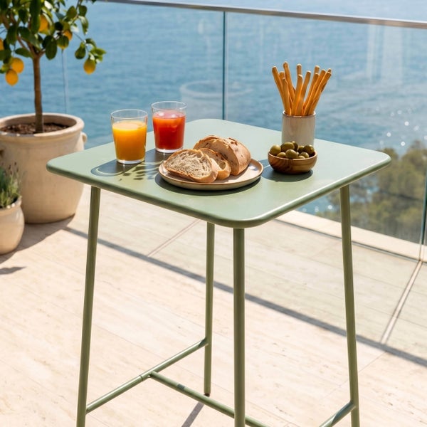 Quadratischer Bistro-Stehtisch aus Metall auf einem sonnigen Balkon mit Meerblick, gedeckt mit Getränken und Snacks.
