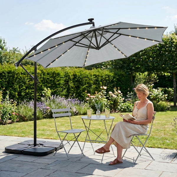 Großer Ampelschirm mit LED-Beleuchtung über einem bistro set aus Metall mit zwei Klappstühlen und Tisch auf einer Terrasse im Garten.